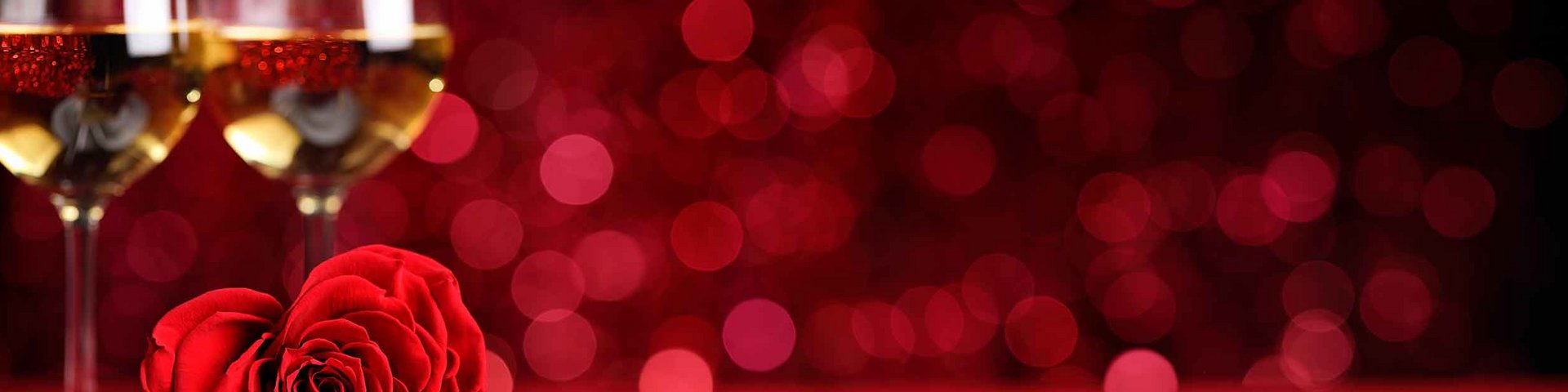 Et hjerteformet rødt rose ligger på en hvid tallerken med bestik ved siden af. I baggrunden ses to glassene fyldt med gylden drink, alt på en rød baggrund. Billedet giver en romantisk stemning.